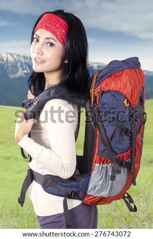 Beautiful woman carrying backpack for hiking, smiling on the camera while walking on the mountain