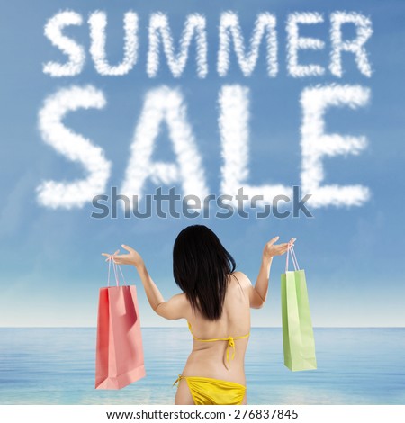 Back view of female shopaholic wearing swimsuit on the shore while holding two shopping bags under cloud shaped a summer sale text