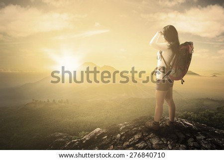 Rear view of young woman standing on the mountain peak while carrying backpack and enjoying sunrise view