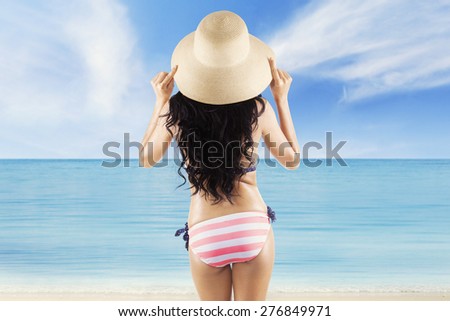 Rear view of sexy woman with long hair standing on the beach while wearing hat, enjoy beach view at summertime