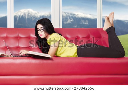 Portrait of young girl lying on sofa while studying with textbooks and laptop computer