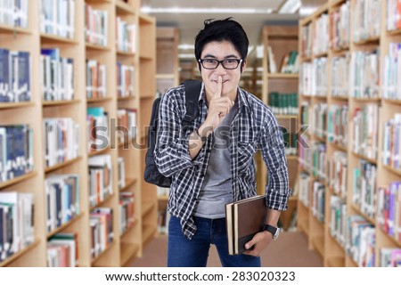 Portrait of male high school student standing in the library and make silence sign
