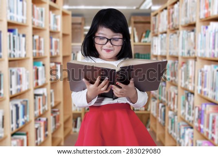 Portrait of beautiful little girl reading a book in the library aisle while wearing glasses