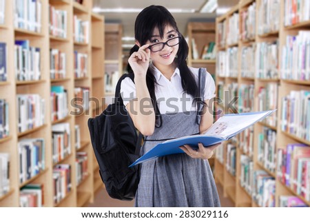 Beautiful young high school student holding a folder in the library while looking at the camera