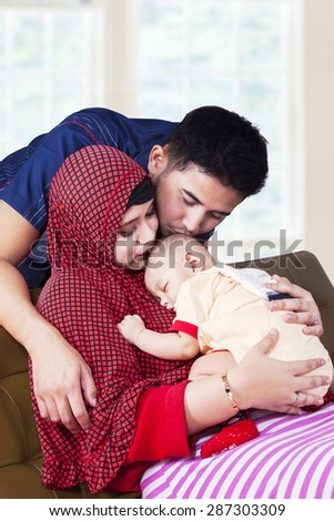Portrait of two young muslim parents kiss their baby boy on the sofa at home