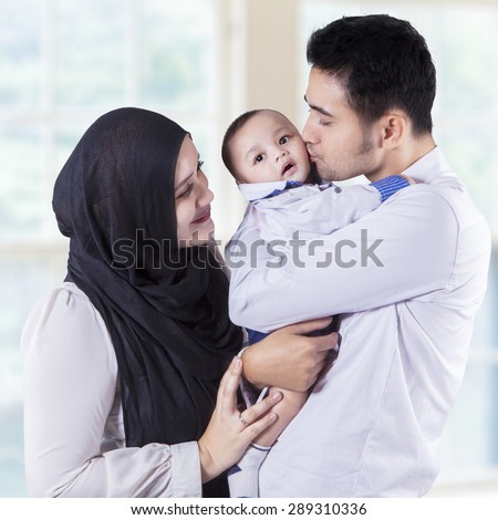 Portrait of happy muslim family kiss their baby while standing near the window at home