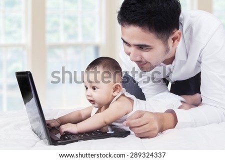 Portrait of joyful infant looks curious with a laptop and try to type, shot with dad at home