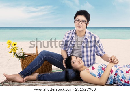 Beautiful asian couple relaxing at beach while sitting on mat and smiling at the camera