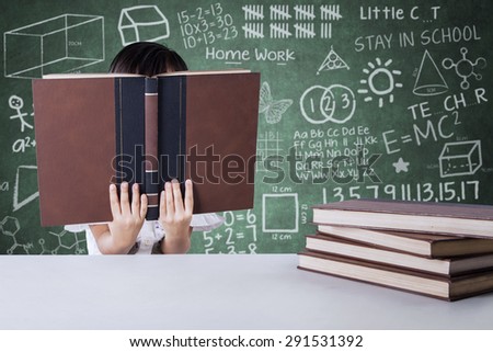 Little girl reading textbooks in the class while covering her face, shot with doodle background on the blackboard