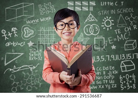 Portrait of pretty little girl standing near the chalkboard in the class while reading a book