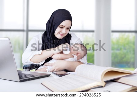 Portrait of young mother feeding her baby with milk from bottle while working with books and laptop