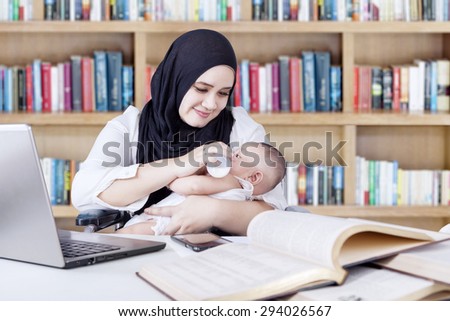 Multitasking young mother nursing her baby with milk from bottle while working in the library