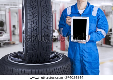 Male mechanic with blue uniform showing empty tablet screen near the tires, shot at workshop