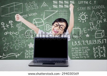 Joyful elementary school student celebrate her success and raise hands in the class with laptop on the table