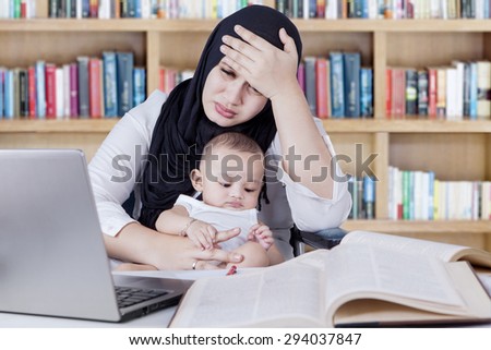 Portrait of frustrated mother carrying her baby while working with laptop and textbooks in library