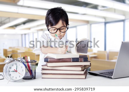 Cute female kindergarten student studying in the classroom and writing on the book with laptop and alarm clock on the table