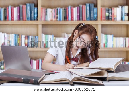 Female student with long hair sitting in the library while writing on the book with laptop at desk