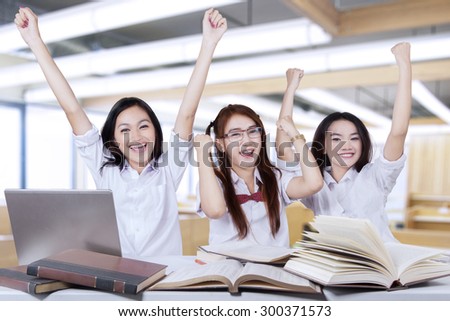 Portrait of three cheerful schoolgirls raise hands together in the classroom while studying with books and laptop