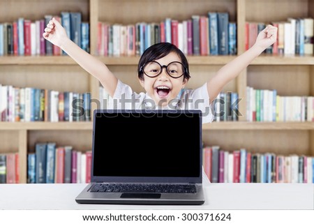 Attractive elementary school student celebrating her achievement in the library with laptop on desk