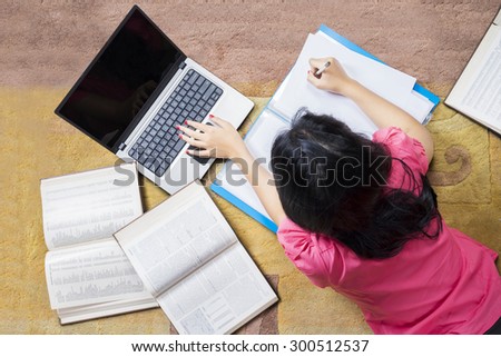 Unique perspective of female high school student studying with laptop and books while lying on the carpet at home