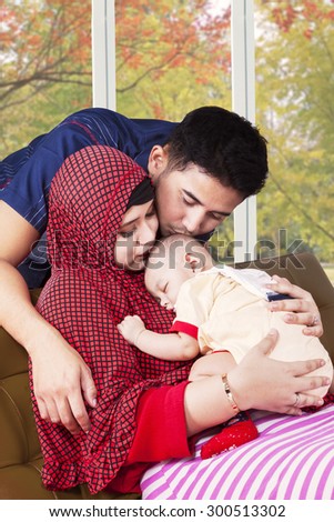 Attractive two young muslim parents kiss their child on the sofa, shot with autumn background on the window