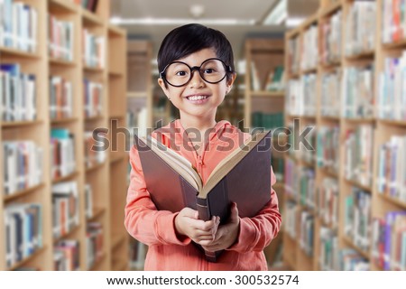 Attractive female elementary school student reading a literature in the library and smiling on the camera