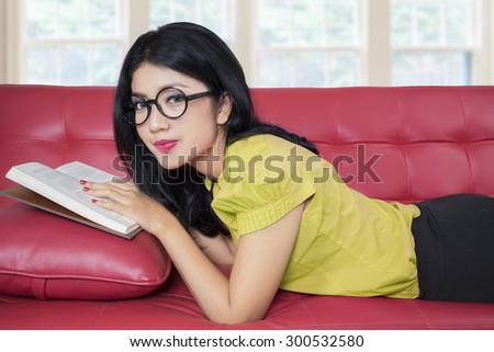 Beautiful young woman smiling on the camera while lying on the couch and reading a book