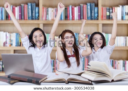 Three beautiful teenage students expressing their success by raising hands together in the library