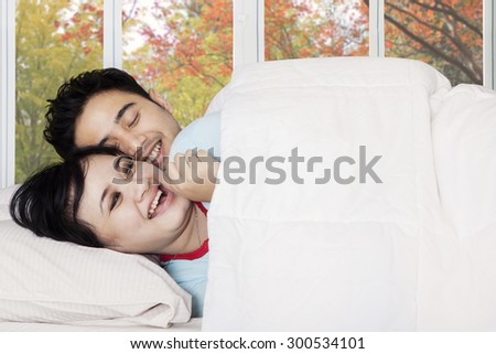 Cheerful young couple lying on bed while joking under blanket with autumn background on the window