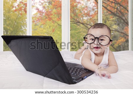 Excited little baby boy lying on bed with laptop computer and wearing a round glasses
