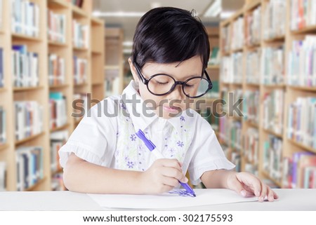 Beautiful little girl wearing glasses in the library and using a marker to draw on a paper