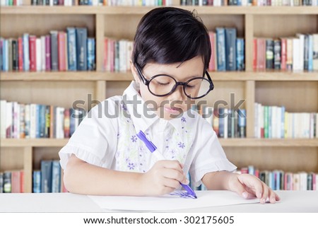 Portrait of little girl sitting in the library while drawing on a paper with a marker and wearing glasses