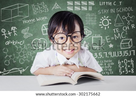 Attractive little student smiling on the camera with a book on the table, shot in the class with scribble background on the chalkboard