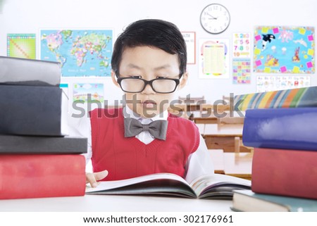 Cute little boy studying in the classroom and reading lesson books while wearing glasses, shot in the school