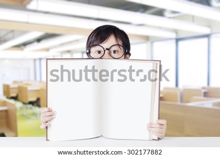 Little kindergarten student sitting in the classroom and showing empty book while wearing glasses