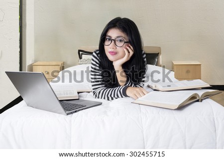 Sweet woman relaxing on bed and smiling on the camera with laptop and books