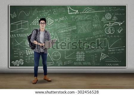 Handsome male high school student standing in class while holding laptop computer