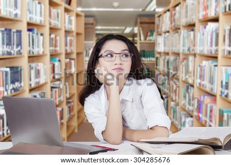 Cute schoolgirl with black and long hair, wearing glasses and daydreaming in the library with books and laptop on desk