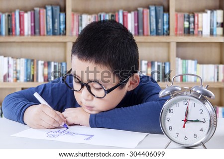 Portrait of child wearing glasses and drawing on a paper in the library with a clock on the table