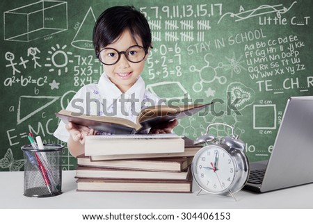 Little schoolgirl reading books in the classroom with alarm clock, laptop and stationery on the table