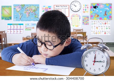 Portrait of attractive schoolboy drawing on the paper in the classroom with a clock on the table