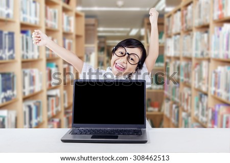 Cheerful little girl raise hands to celebrate her success in the library with empty laptop screen on the table