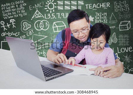 Portrait of female elementary school student studying in the classroom with her teacher, using a book and laptop on the table