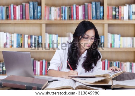 Teenage girl with long hair sitting in the library while studying and doing her school assignment with books and laptop