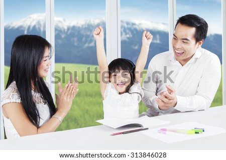 Photo of two young parents giving applause on their daughter after finishing school assignment at home
