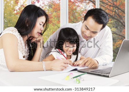 Photo of attractive little girl try to write on the book with her parents at home, shot with autumn background on the window
