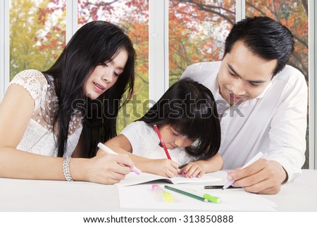 Image of a cute and smart girl with black hair doing school assignment at home with her father and mother