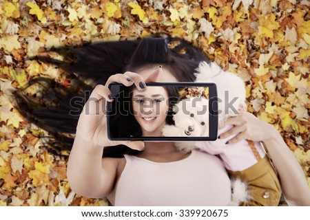 Unique perspective of beautiful woman using smartphone to take selfie photo with her dog on the autumn leaves