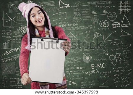 Joyful female student wearing warm clothes in the class and showing blank clipboard