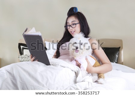 Young woman holding book and reading it while resting on bed with her dog